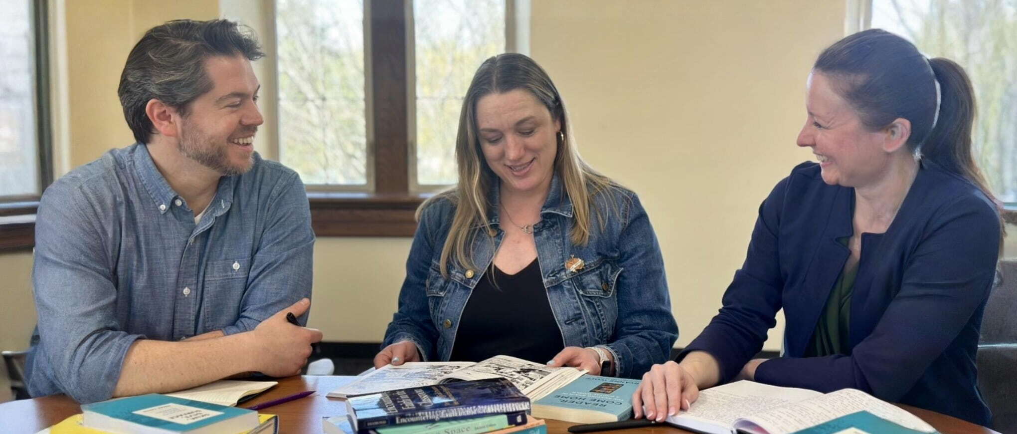 Three people smiling and looking at books and papers around a wooden table.