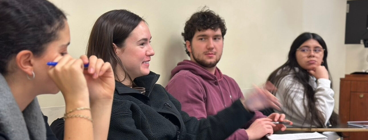 Four students at desks. A woman in black fleece smiles, gesturing. A man in maroon hoodie looks right, another woman rests her chin.
